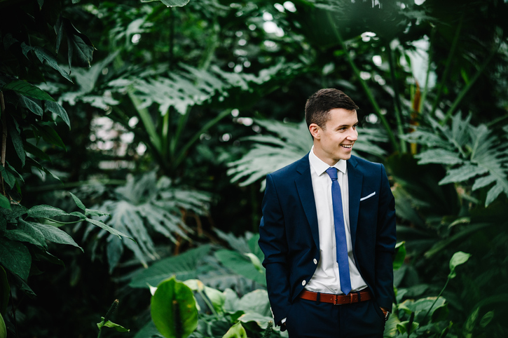 Happy portrait handsome groom in a wedding suit and tie is standing on the background of greenery. Man stands in the Botanical garden full of greenery.